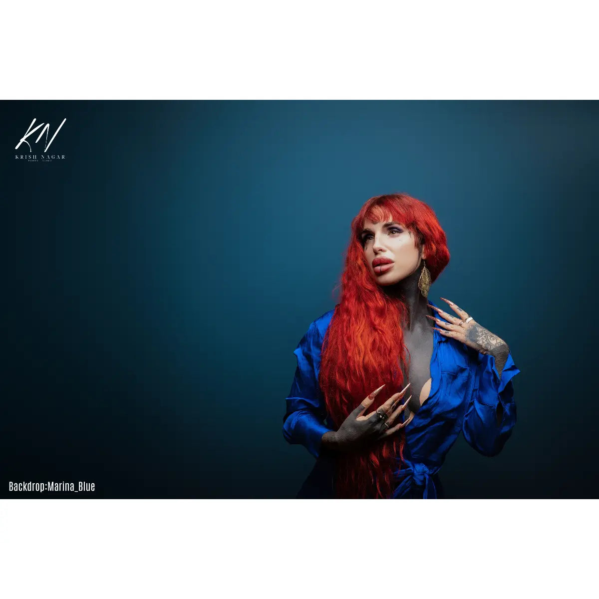 Woman with red hair and tattoos wearing a blue outfit against a dark blue backdrop