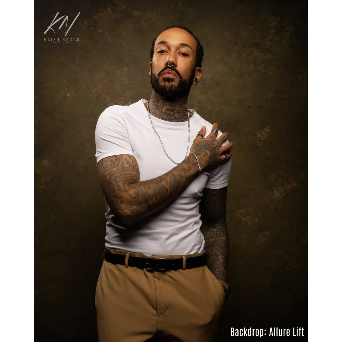 Man with tattoos wearing a white t-shirt and brown pants against a dark background