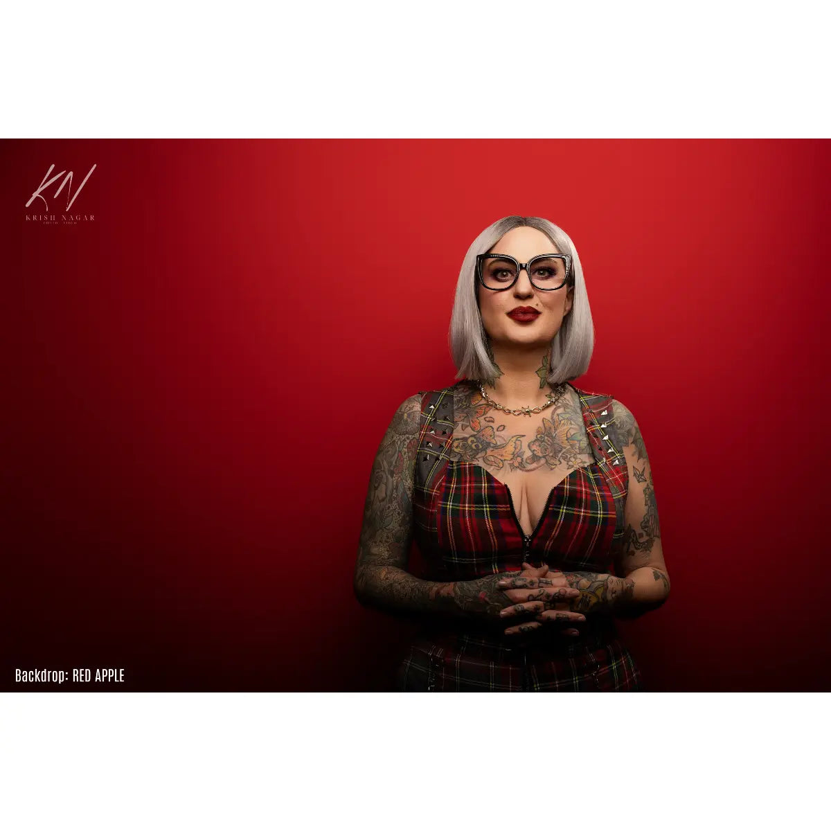 Woman with tattoos and glasses wearing a plaid dress against a red background