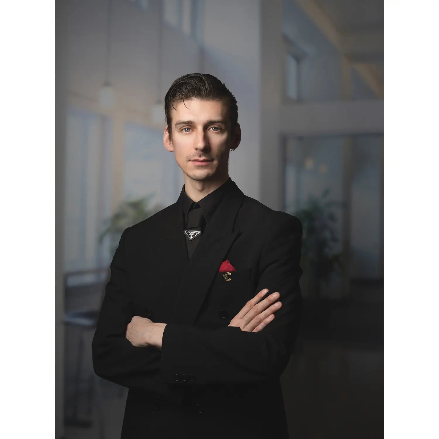 Man in a black suit with a red pocket square standing in an indoor setting.