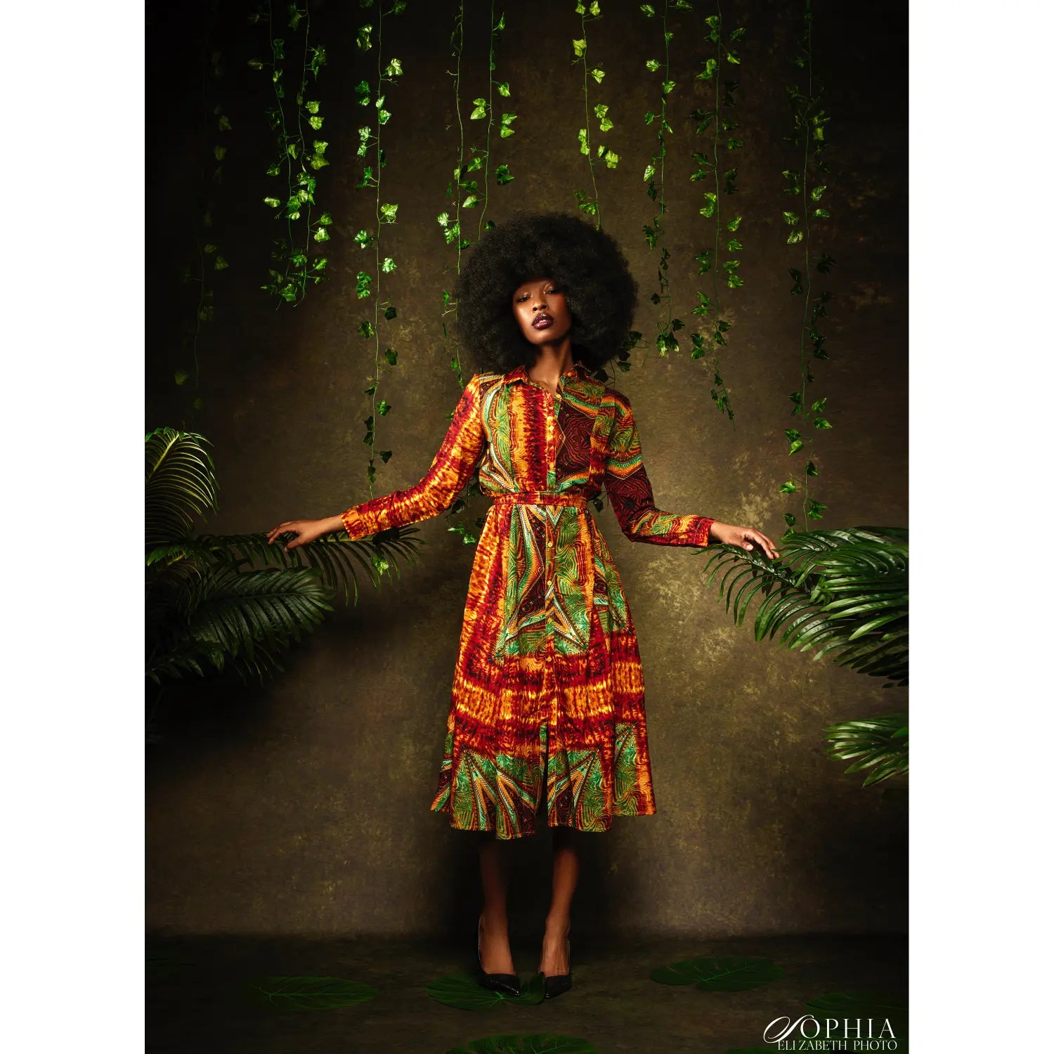 Woman in a colorful dress standing among green plants with a dark background