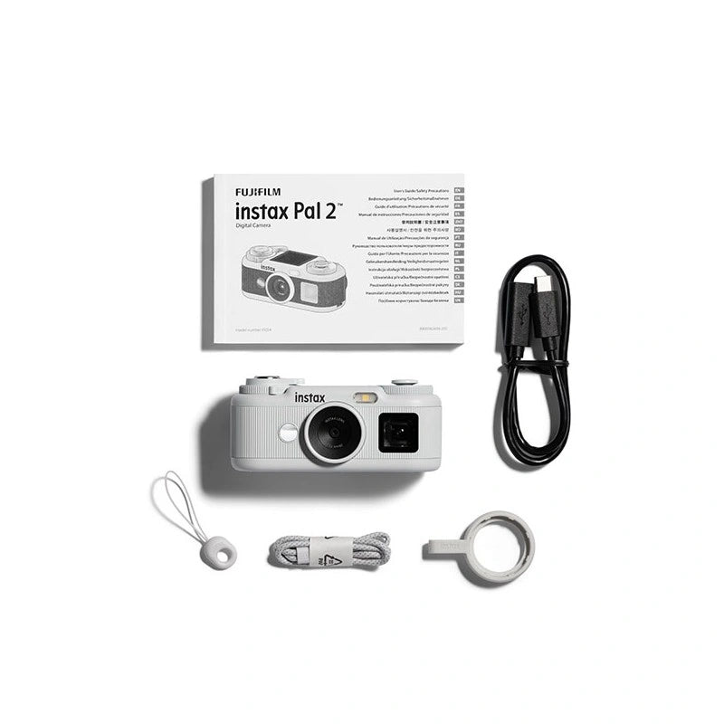 A white Fujifilm Instax Pal 2 digital camera shown with its retail box, black USB cable, wrist strap, charging cable, and MagSafe ring on a clean white background.