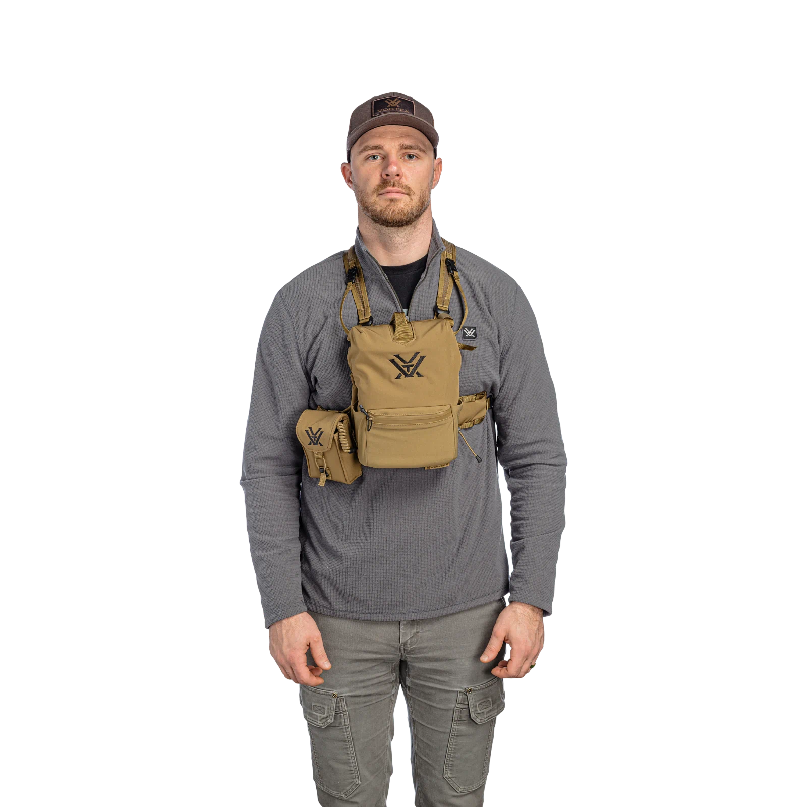 A man wearing a gray long-sleeve shirt and gray cargo pants models a tan harness vest with a side pouch, featuring the Vortex logo, against a solid white background.
