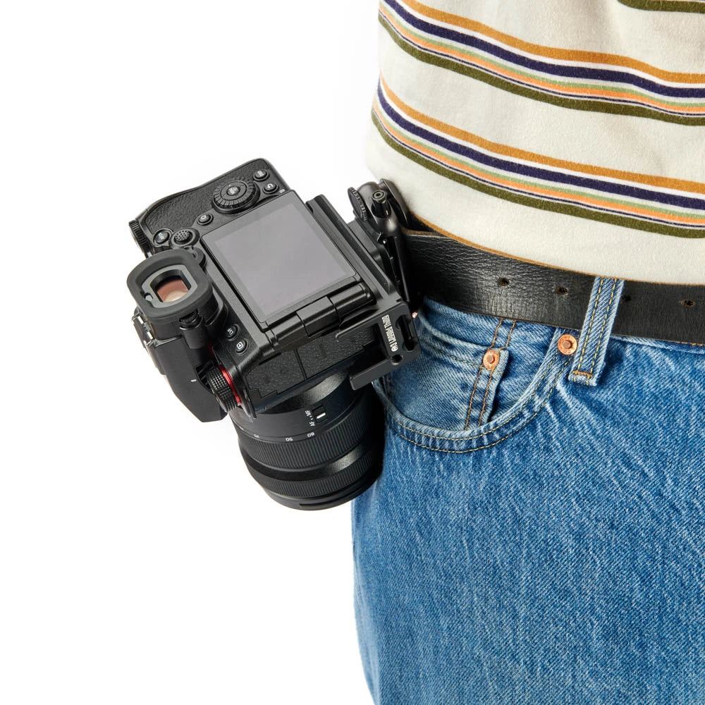 A black mirrorless camera with an attached lens clipped to a person's belt pocket, shown against a white background. The person is wearing a striped shirt and blue jeans.