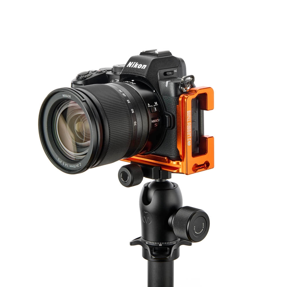 An orange and black camera mounting bracket attached to a Nikon mirrorless camera with a lens, displayed on a black tripod against a solid white background.