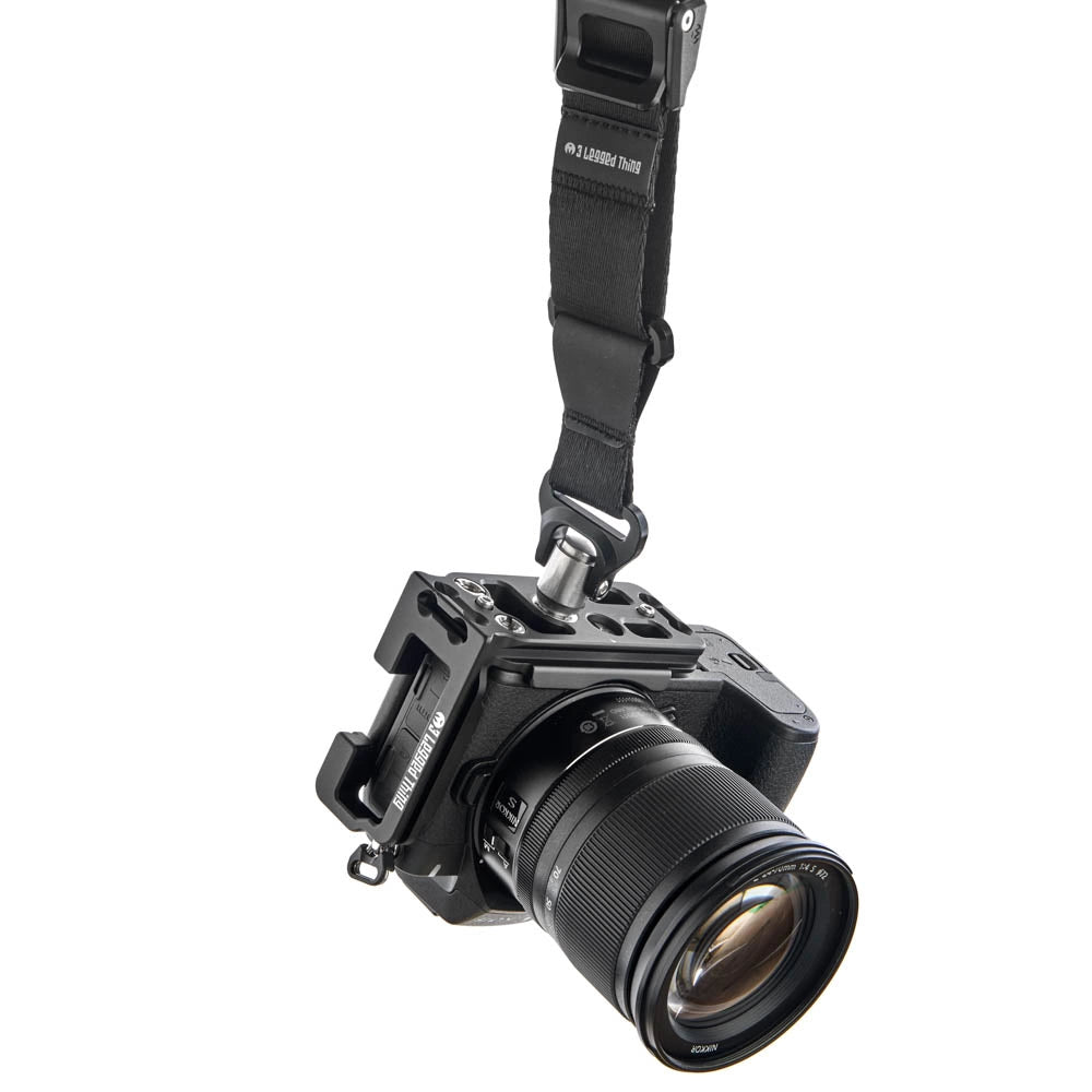 A black camera body with an attached lens, shown hanging from a black camera strap against a plain white background.