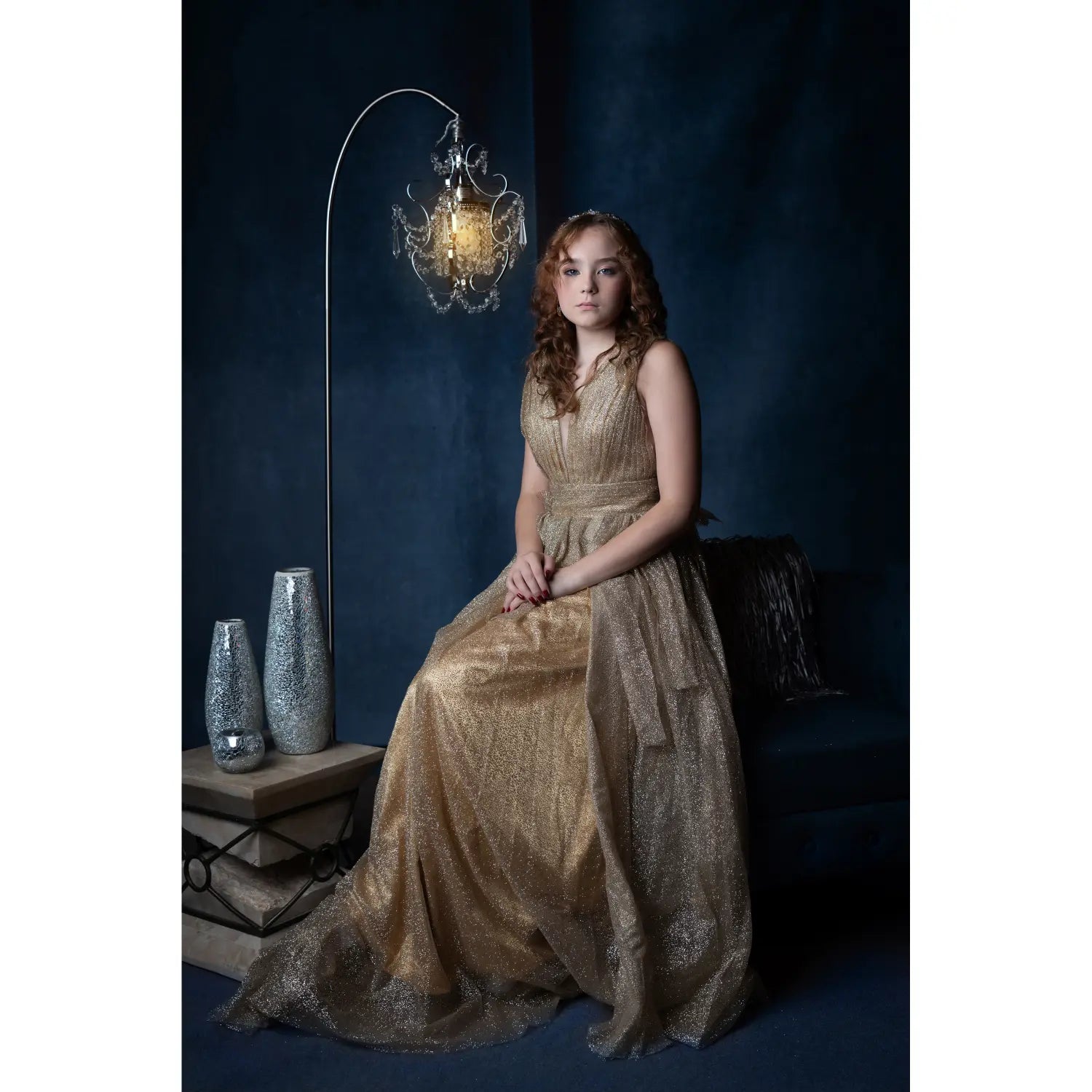 Woman in a gold dress sitting in a dark room with decorative vases and a chandelier.