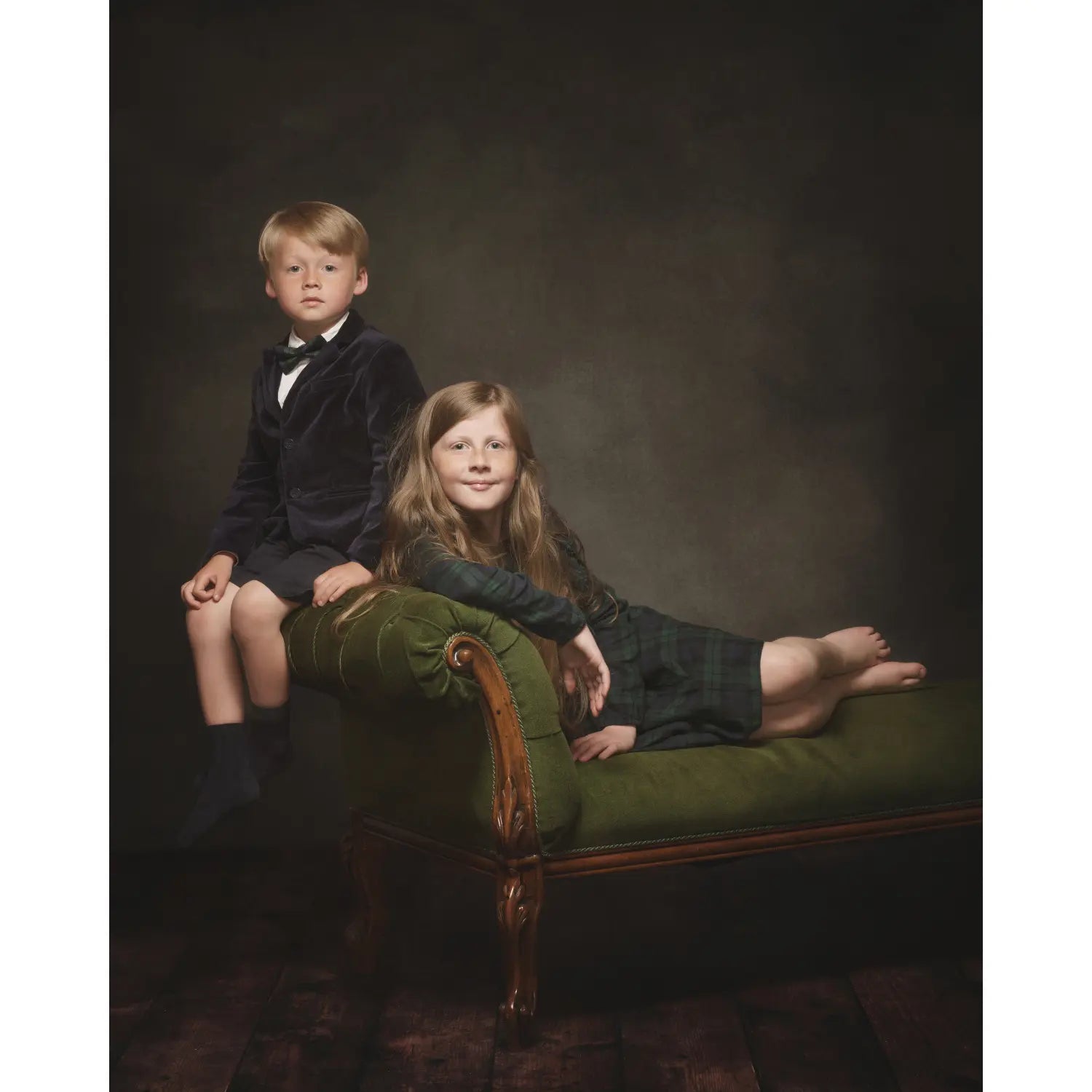 Two children in formal attire sitting on a green velvet couch against a dark background