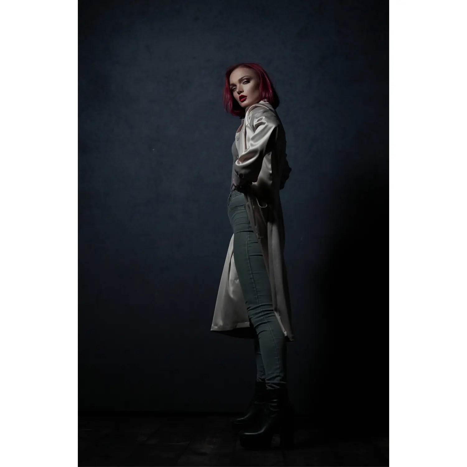 Person with red hair wearing a long beige coat and green pants against a dark background