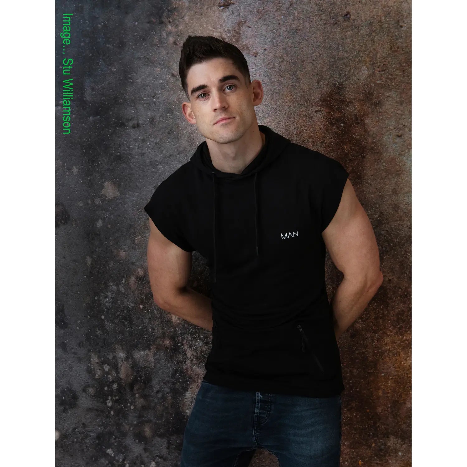 Man wearing a black sleeveless hoodie with a logo against a textured wall.