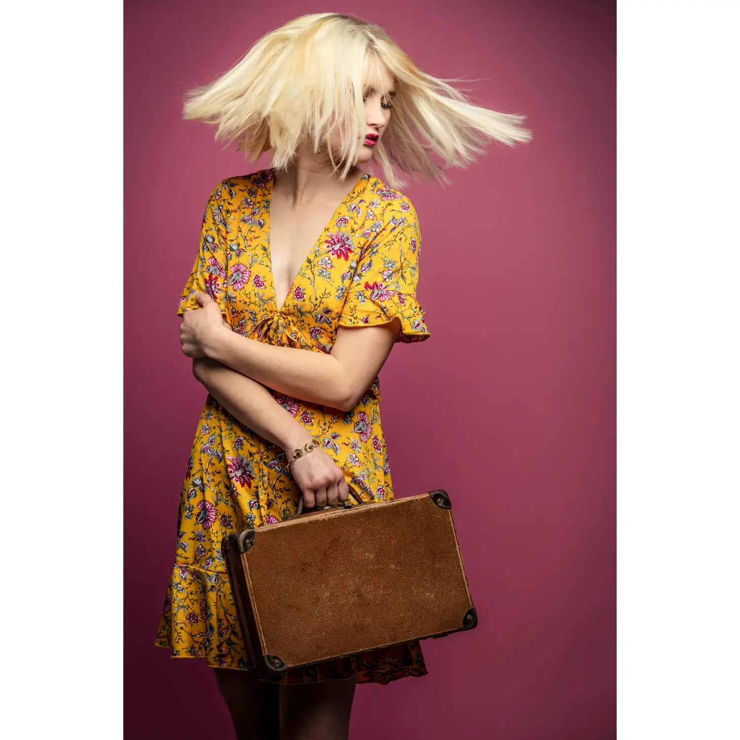 Woman in a yellow floral dress holding a brown suitcase against a purple background