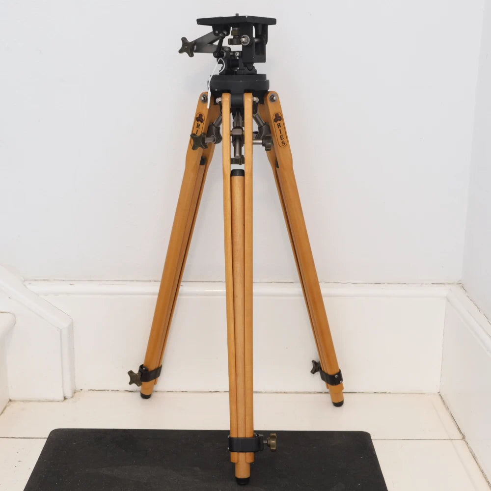 A wooden tripod with a black metal head and legs, standing on a black mat against a white wall and tiled floor.