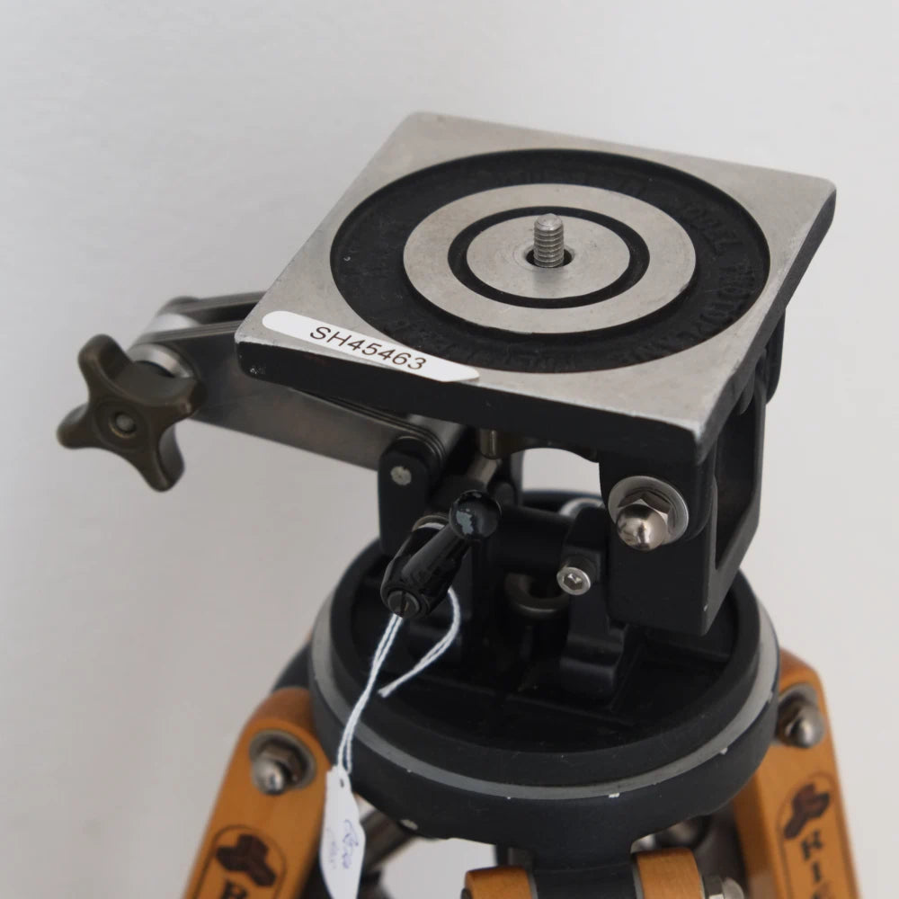 A close-up of a surveying or leveling tripod mount with a square metal target plate featuring concentric black rings, mounted on a black and orange tripod against a plain light background.