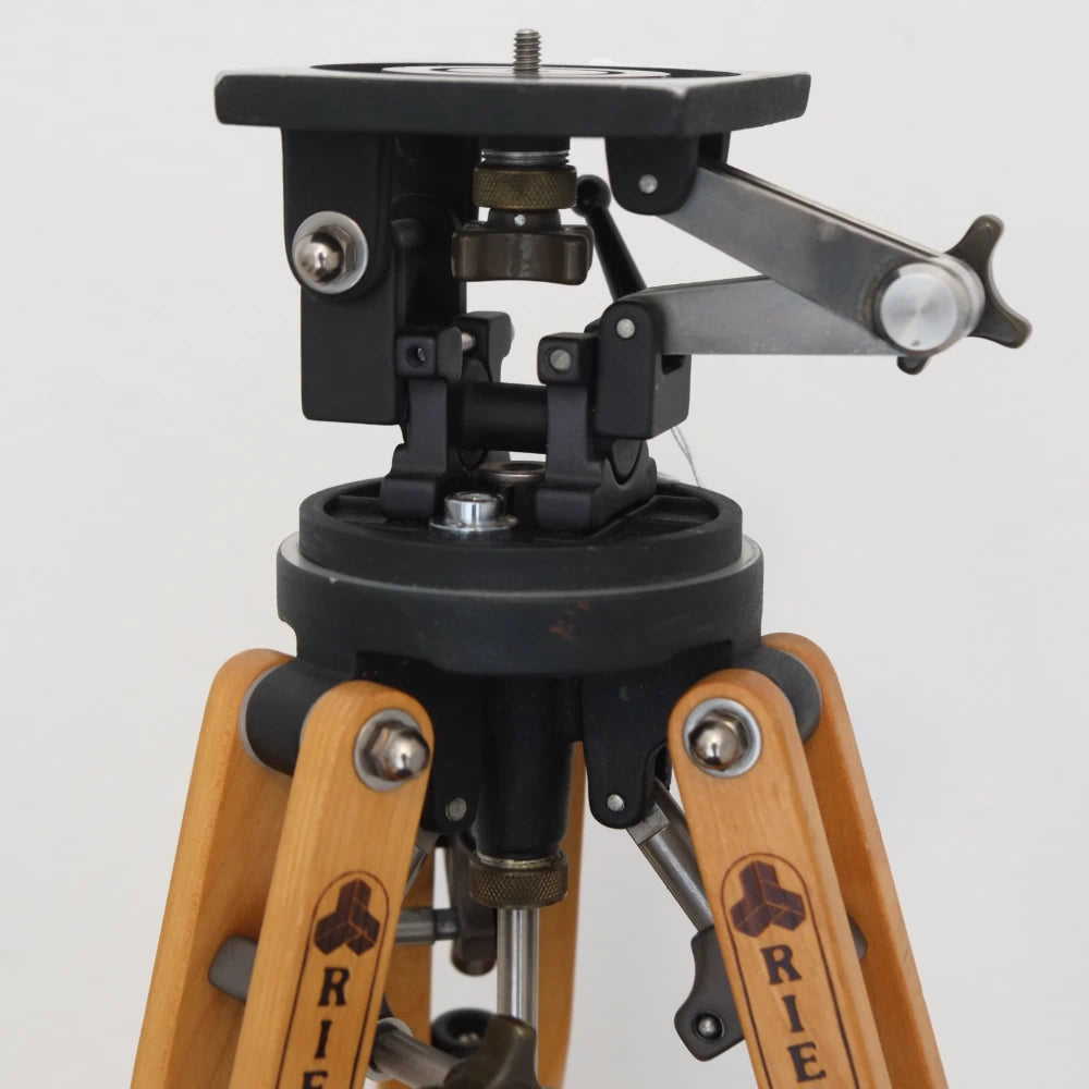 Close-up of a tripod head and lower tripod section with black metal hardware and light wood legs branded RIE, photographed against a plain light background.