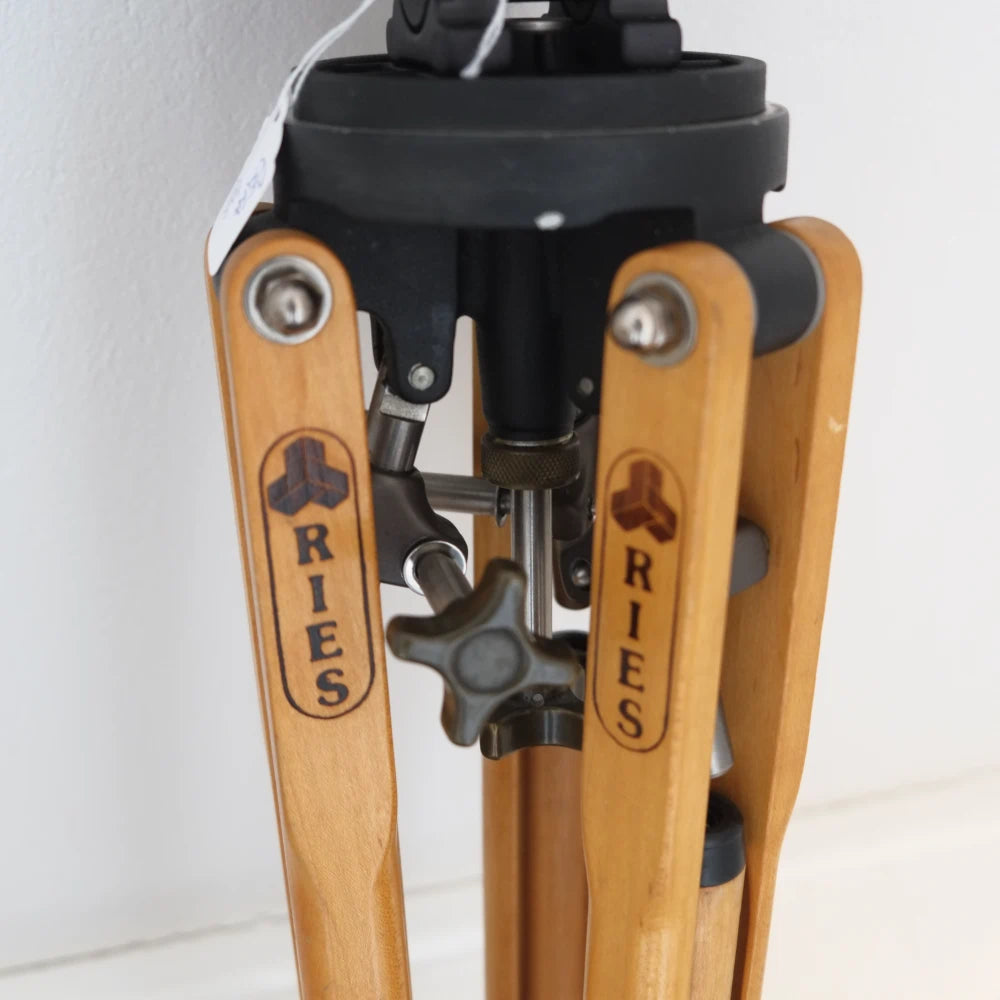 Close-up of a RIES tripod leg with a light natural wood finish and engraved brand logo, showing the metal joint mechanism and black top mount against a plain light background.