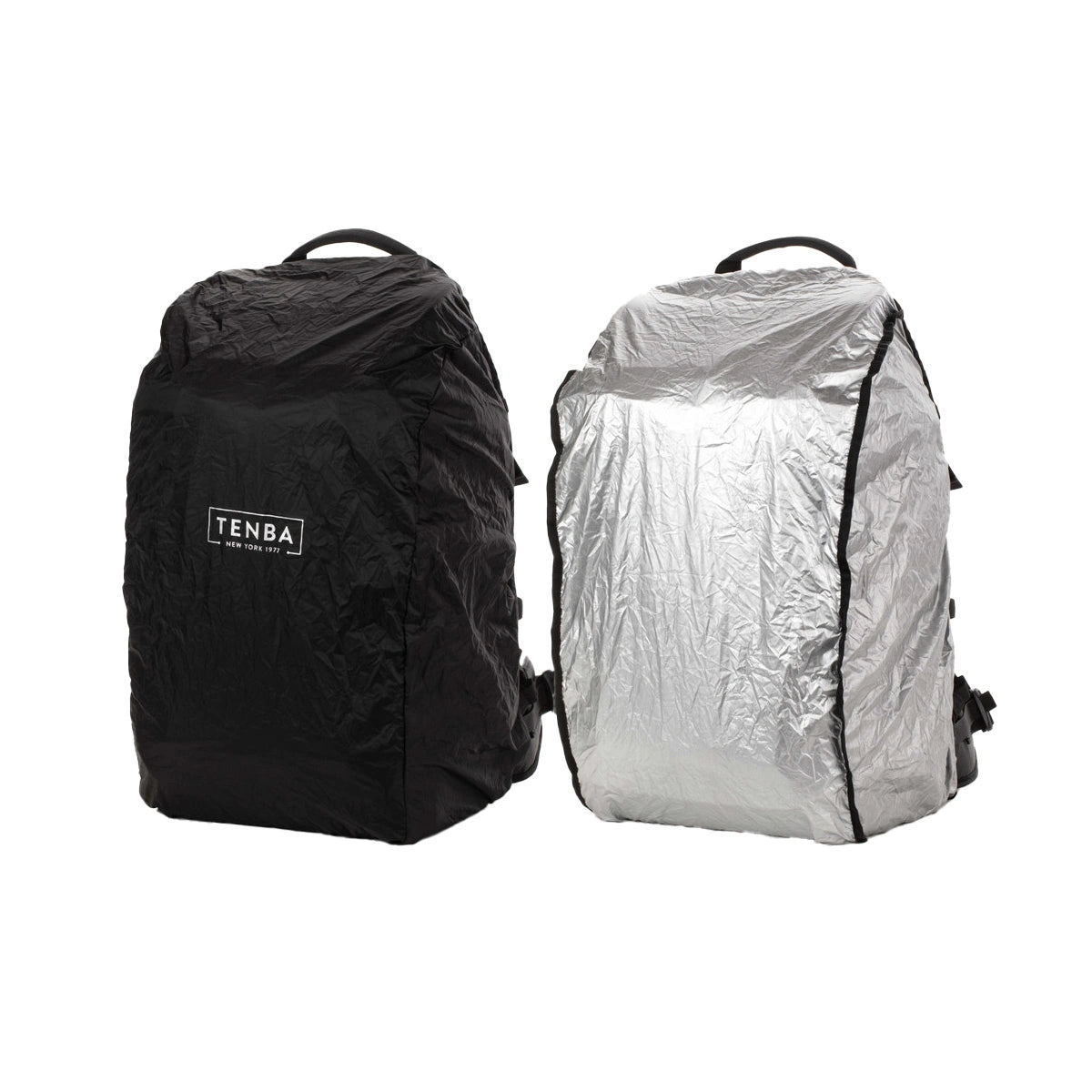 Two backpacks shown side by side on a white background, each covered with a crinkled rain cover. The left cover is black with a visible TENBA logo, and the right cover is metallic silver.
