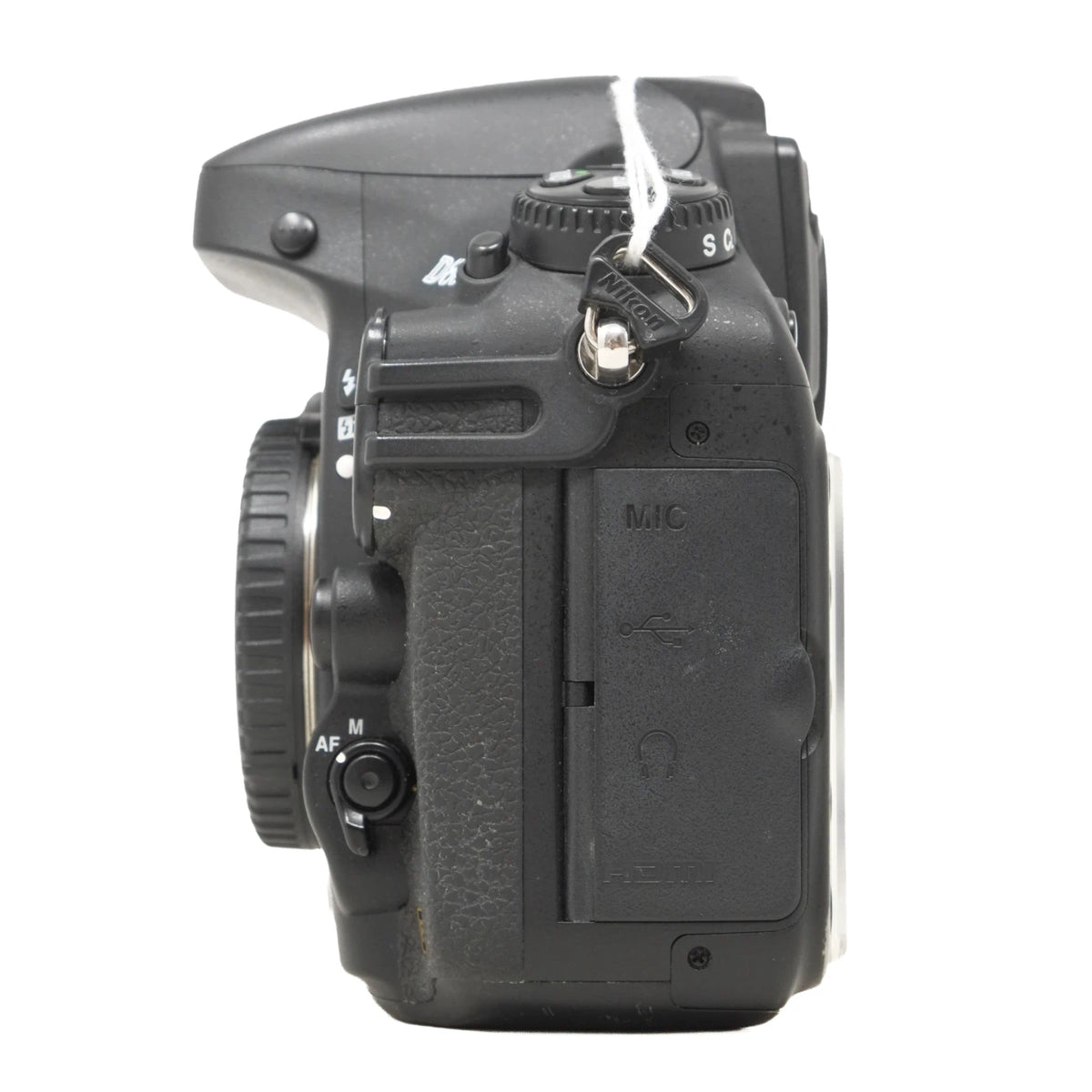 A black Nikon DSLR camera body, viewed from the rear-top angle, showing the mode dial, shutter button, lens mount, and battery compartment cover, with a white string tag attached.
