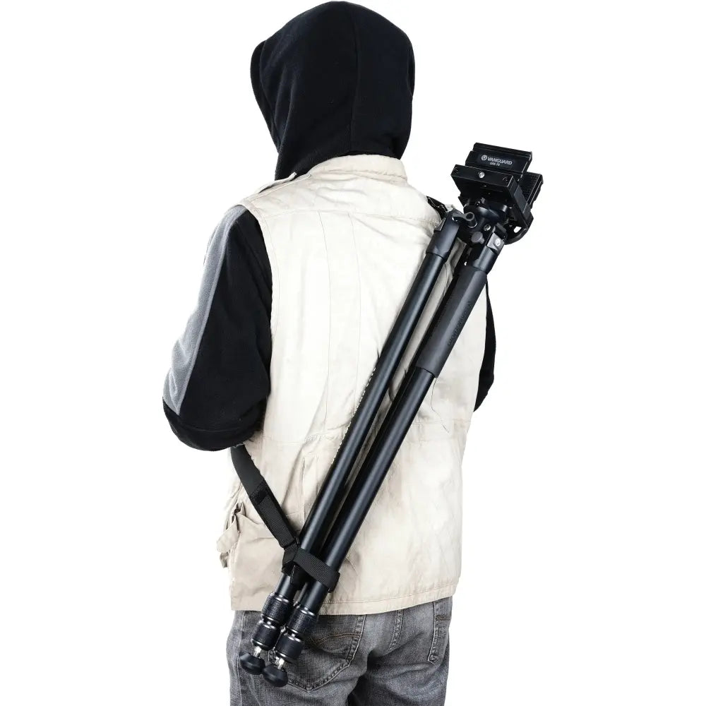 Person wearing a black hooded jacket and beige vest holding a camera tripod on a white background