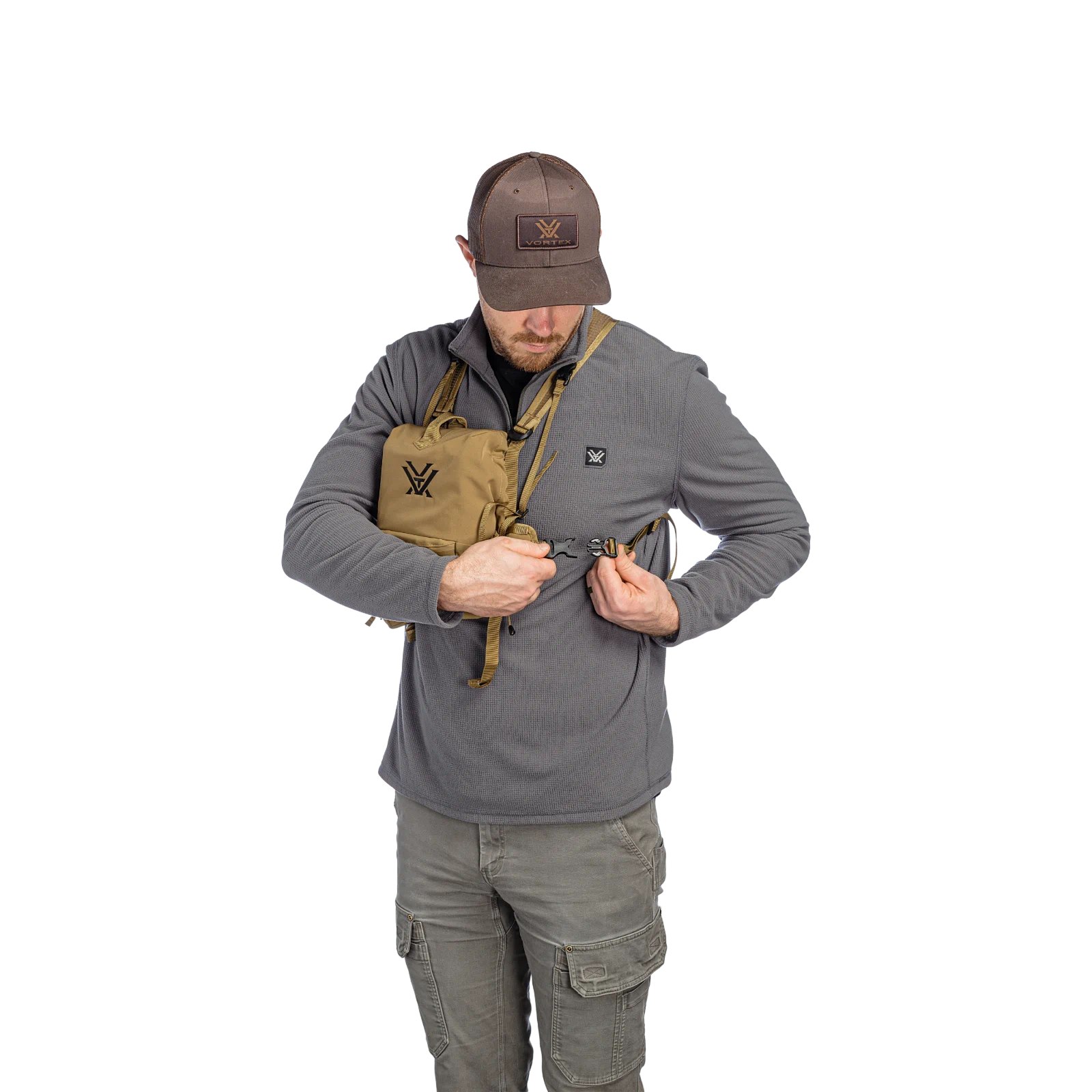 A man wearing a gray long-sleeve shirt, brown cap, and gray cargo pants demonstrates a tan harness with a black logo, shown against a plain white background.
