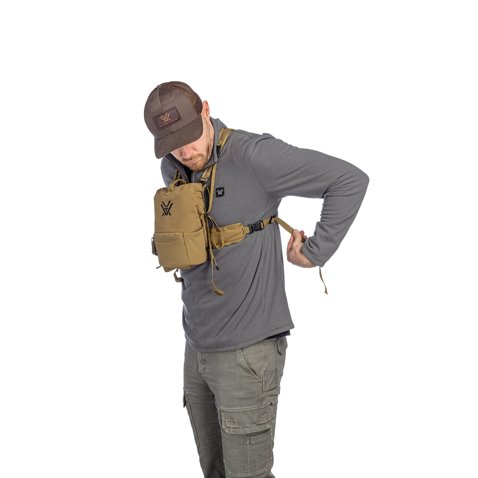 A man wearing a gray long-sleeve shirt, olive cargo pants, and a brown baseball cap models a tan tactical harness with a black logo, shown against a plain white background.